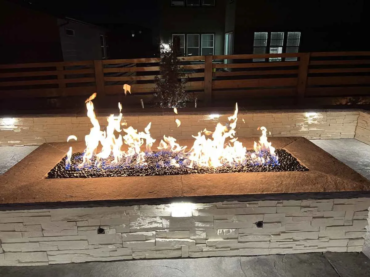 Active linear flame in a rectangular built-in fire pit with dark fire media and stone veneer