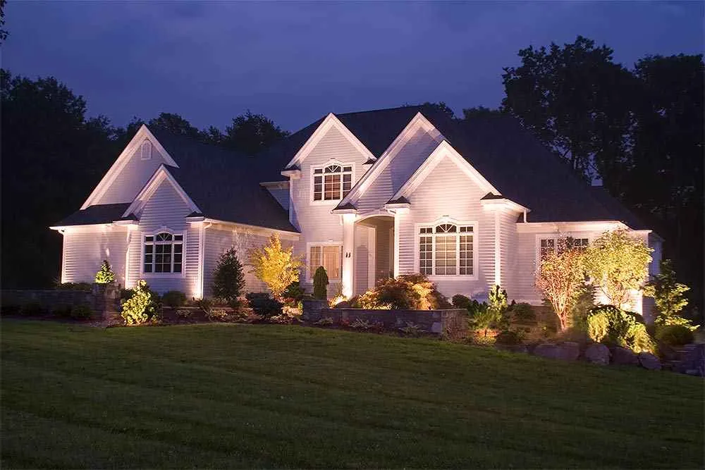 Front entry, trees, shrubs, and a low wall illuminated across a white facade at night