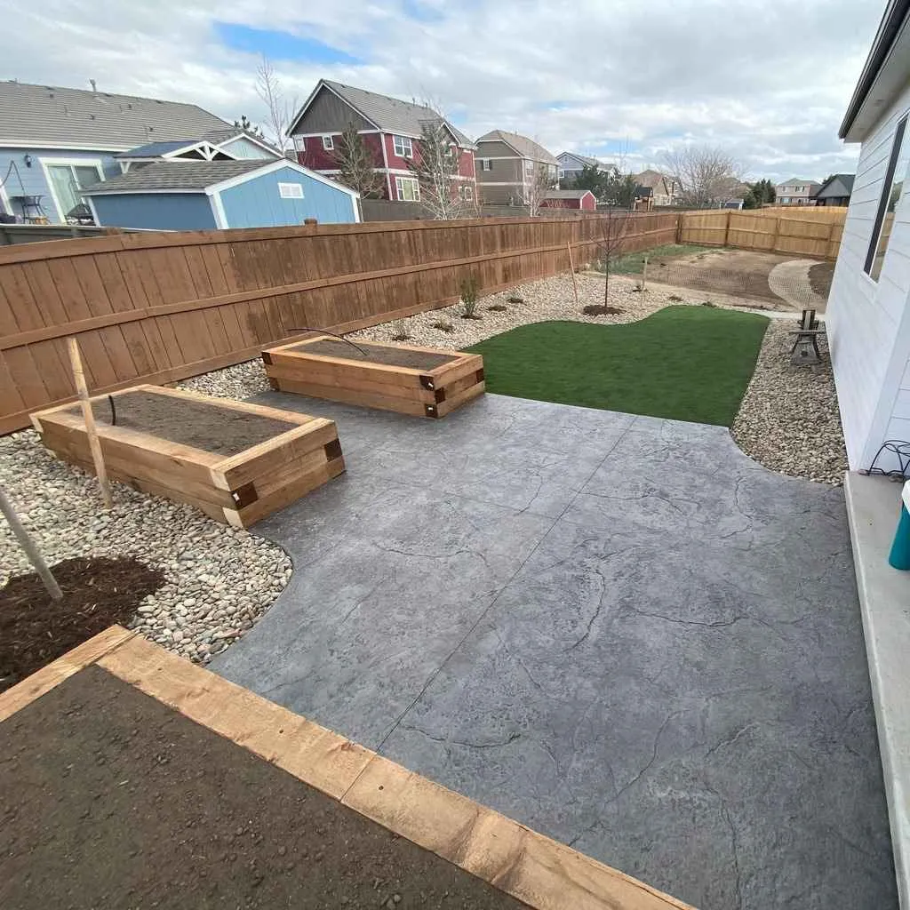 Stamped concrete patio beside raised wood planters, artificial turf, rock, and young trees