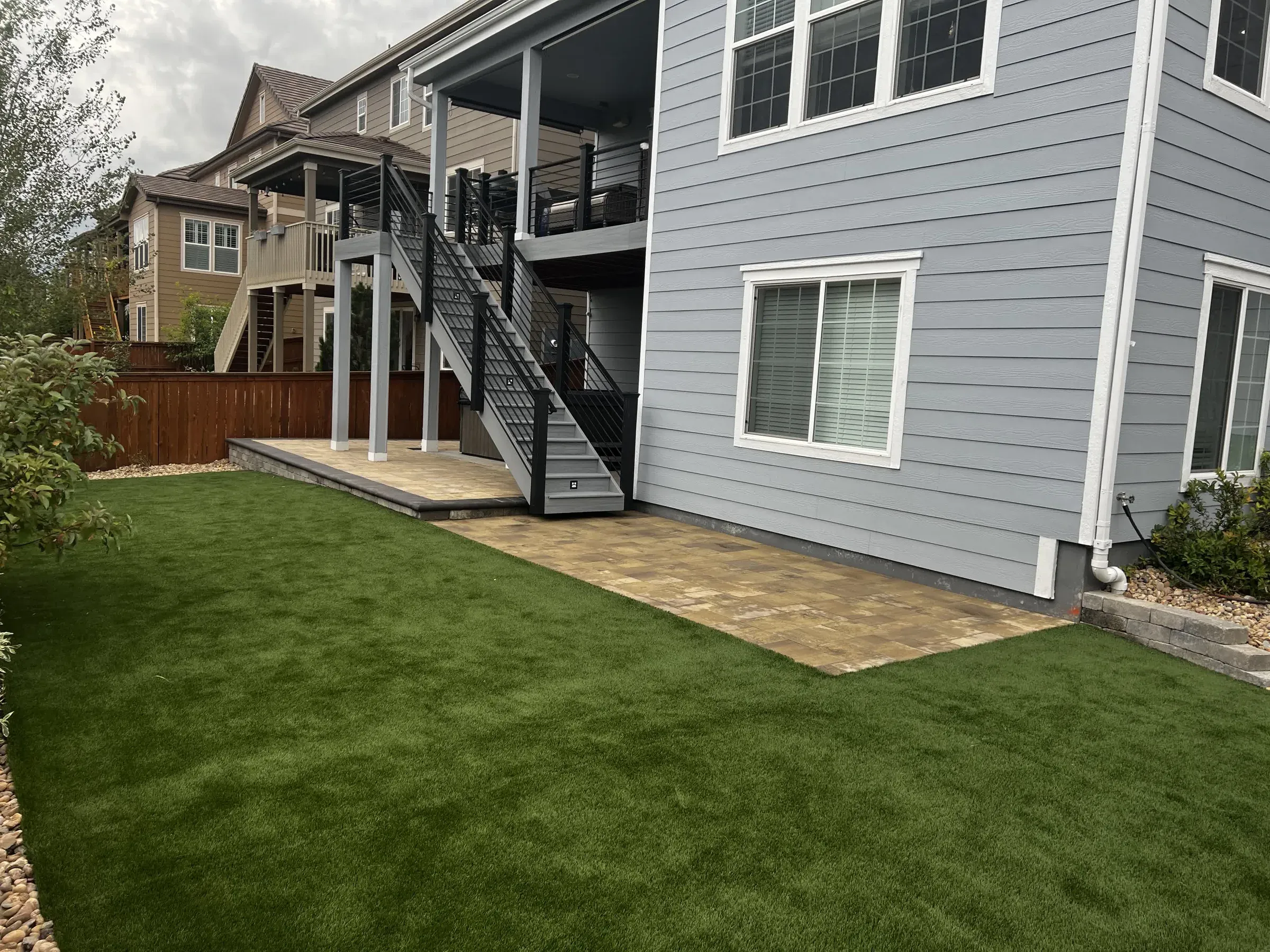 Paver patio beneath a two-story deck stairway next to a backyard artificial turf lawn in Parker